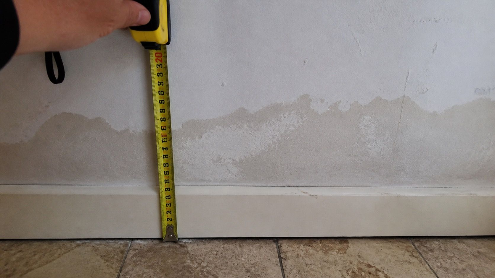 Bas de mur taché, mètre ruban tenu à la verticale contre la plinthe pour donner la hauteur du front d'humidité