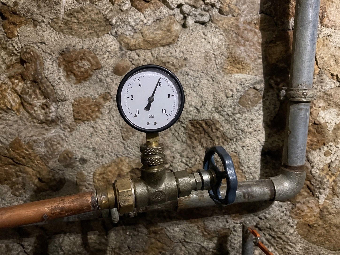 Manomètre raccordé sur une vanne d'arrêt en cave de villa ancienne, aiguille lisible et mur de meulière apparent derrière